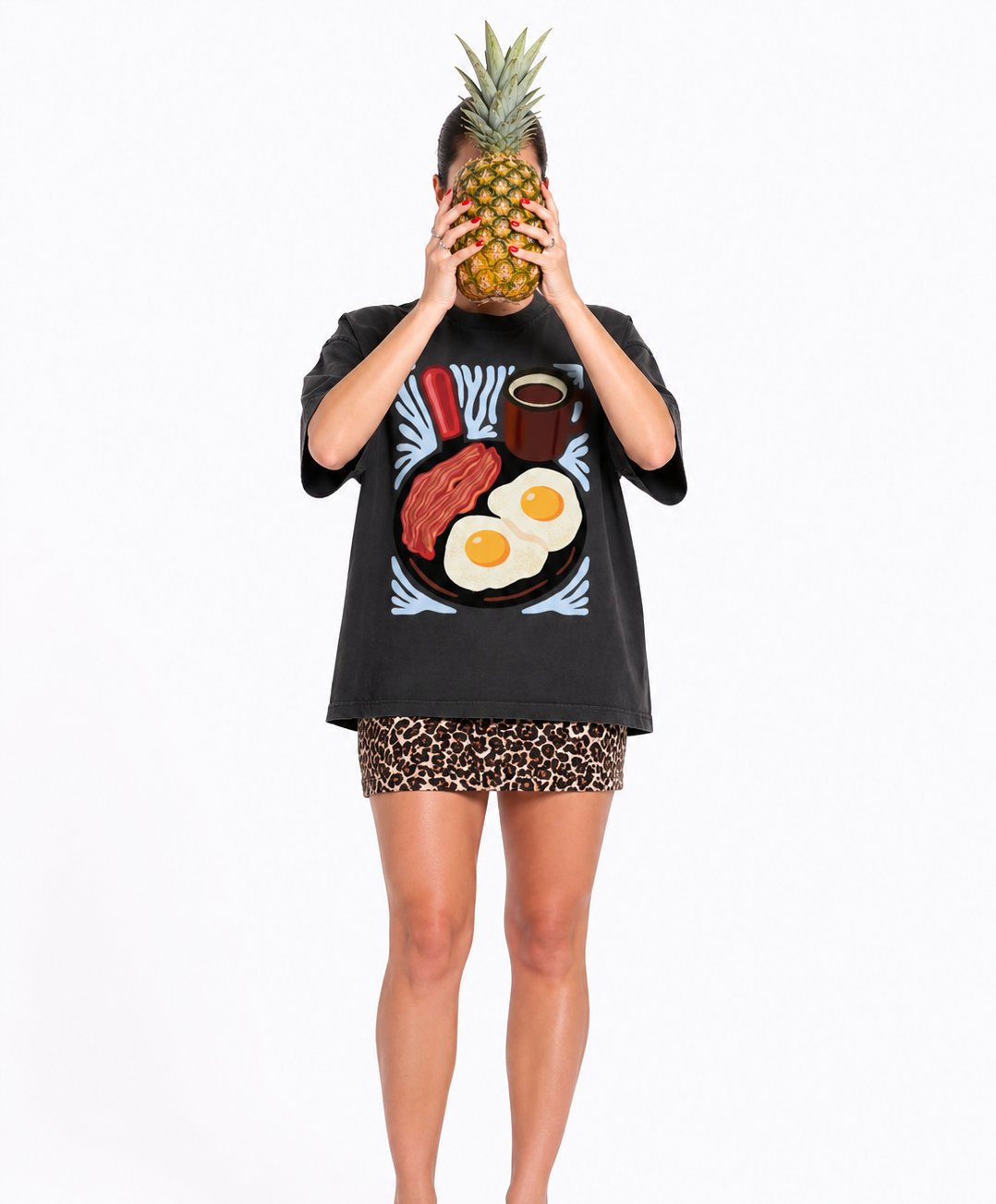 Person wearing a black t-shirt with a graphic design of bacon, eggs, and coffee, holding a pineapple over their head on a white background.