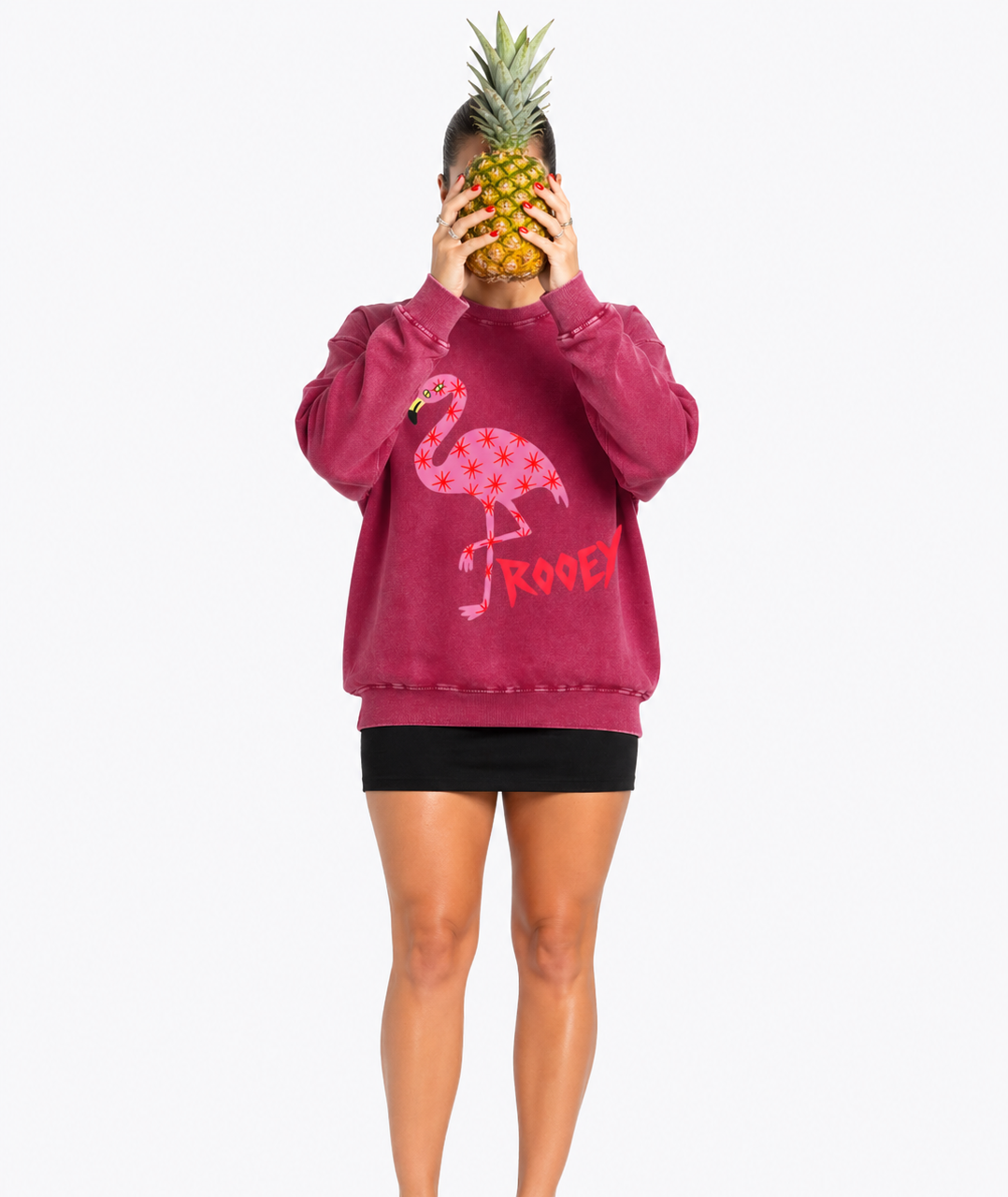 Person wearing a maroon sweatshirt with a flamingo design, holding a pineapple in front of their face on a white background.