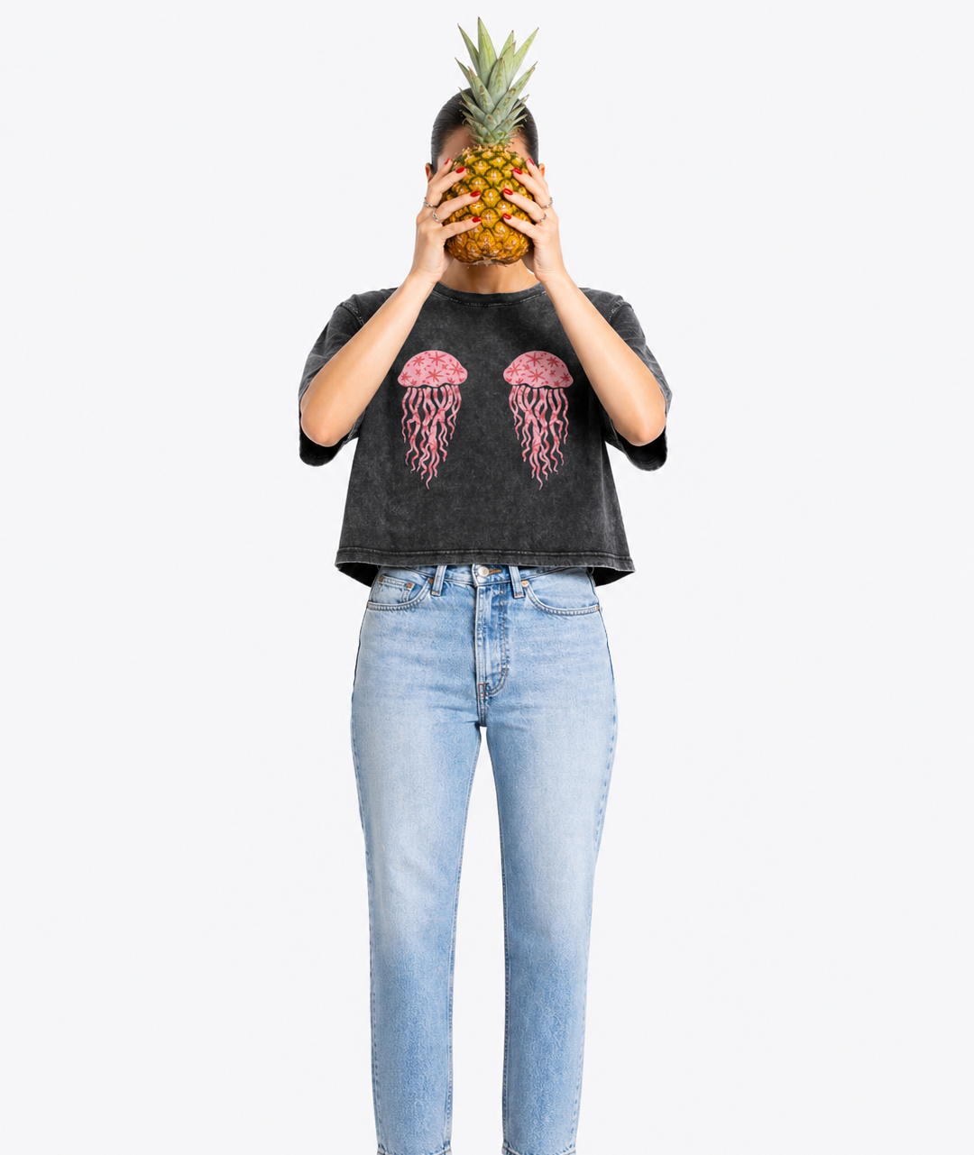 Person wearing a dark gray t-shirt with pink jellyfish design and light blue jeans, holding a pineapple in front of their face on a white background.
