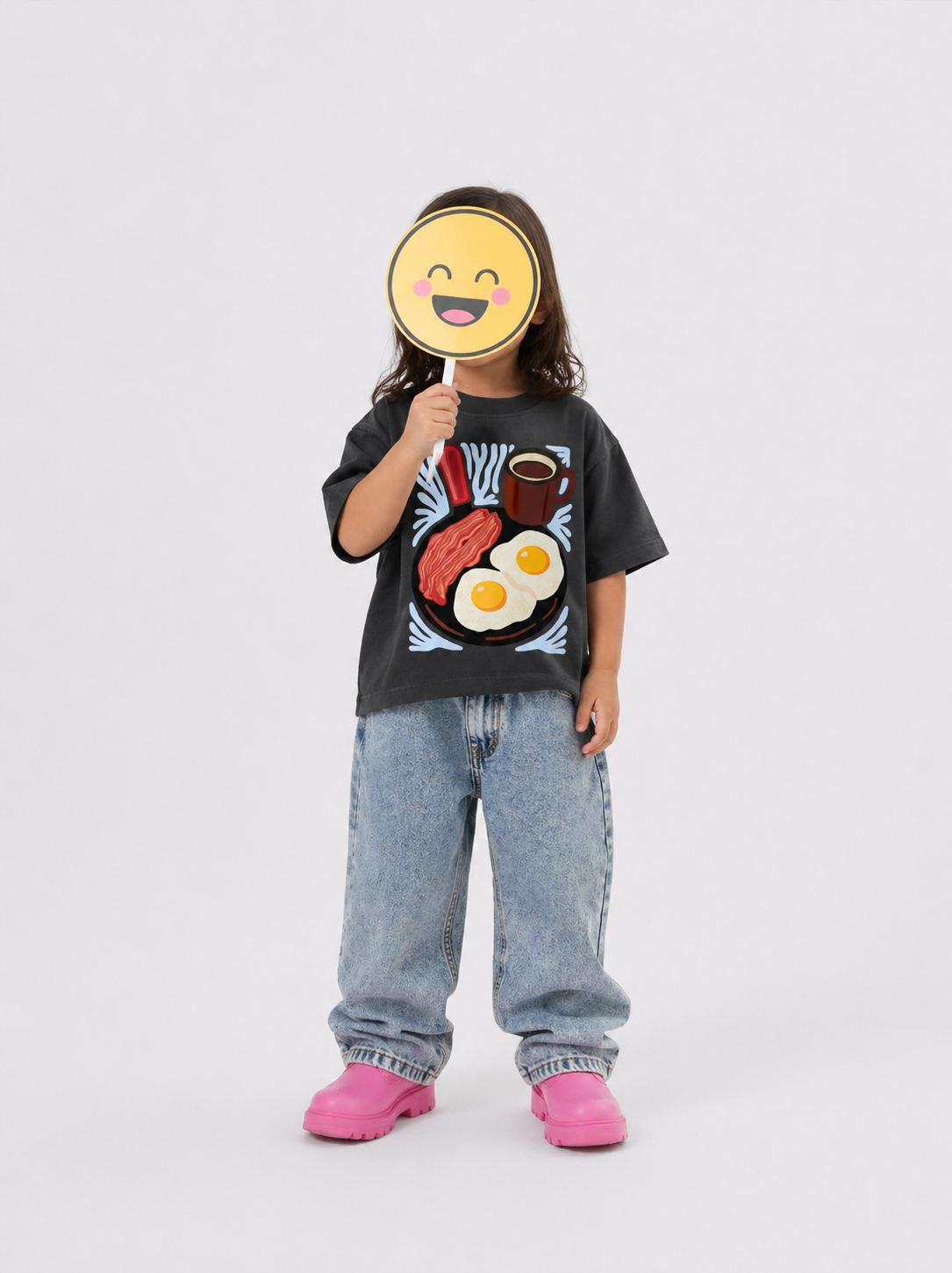 Child wearing a black t-shirt with a graphic design, holding a smiley face mask.